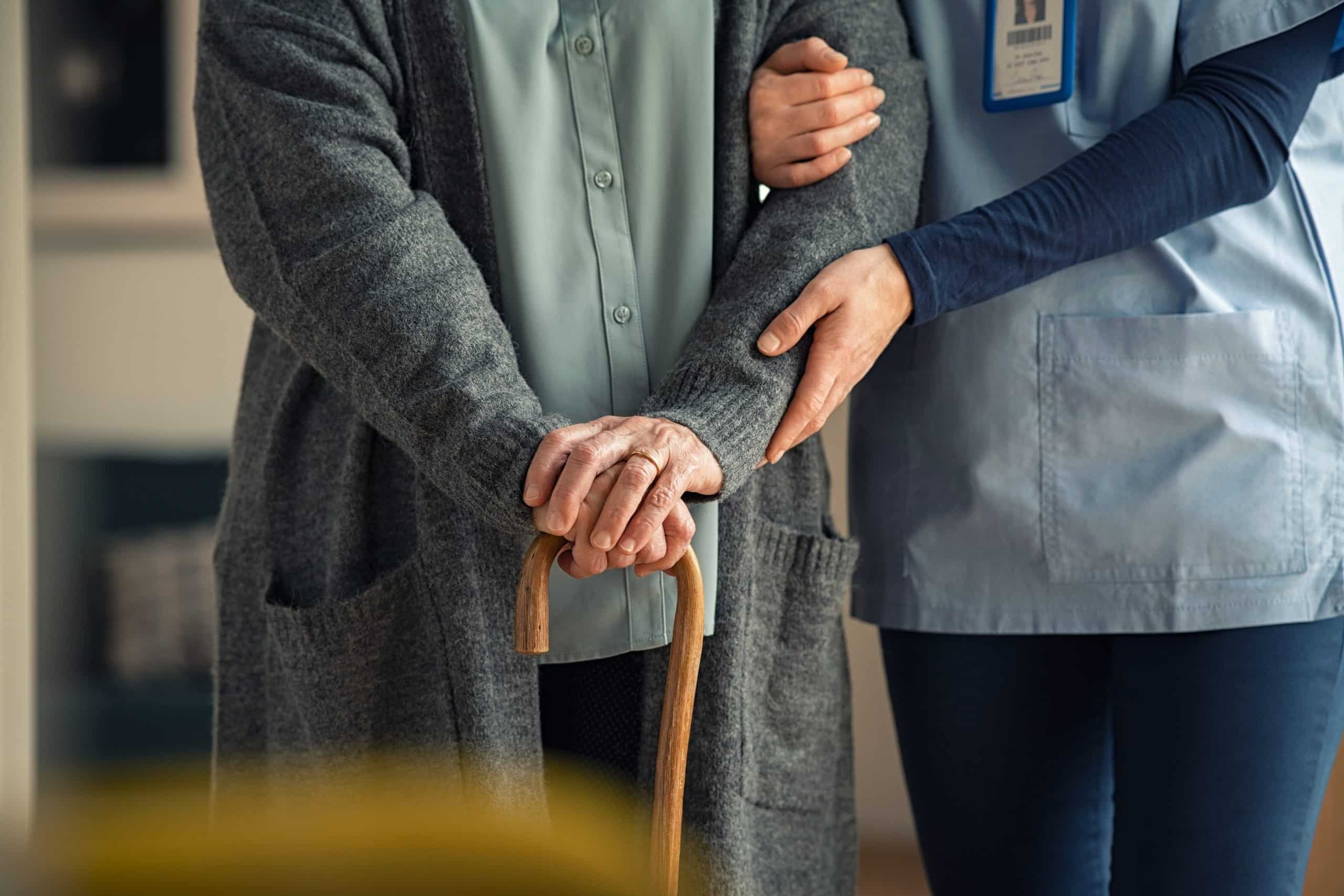 An elderly person holding a cane is being assisted by a caregiver who is holding their arm; both are standing indoors, highlighting compassionate care like that seen in the Bayada Nurse Wage Settlement_Philadelphia.