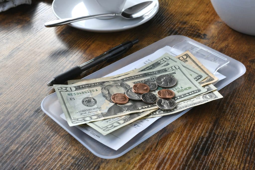 A restaurant bill tray with U.S. dollar bills, coins, a receipt, and a black pen on a wooden table. Other restaurant owners in Philadelphia believe international tourists will be aware of U.S. tipping culture during the games.
